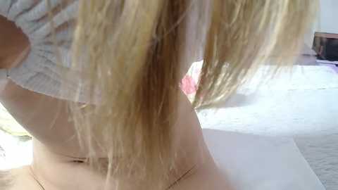 Media: A close-up video of a blonde woman lifting a white sweater, revealing her bare midriff, with a pink bra strap visible. She's indoors, on a white bed, with natural light.