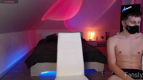 Media: A video of a shirtless, fair-skinned man with a face mask and black shorts sitting on a modern white chair in a dimly lit, minimalist bedroom with a black bed, pink and blue lights, and a nightstand lamp.