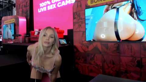 Media: Video of a blonde woman in a pink bra, leaning forward with a playful expression, in front of a red brick wall with a large inflatable boob advertisement.