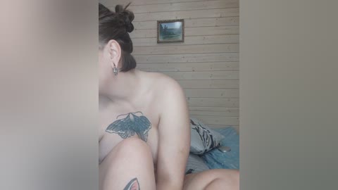 Video of a pale-skinned woman with dark hair in a messy bun, topless, showcasing a large butterfly tattoo on her left shoulder, sitting on a bed in a wood-paneled room with a framed landscape painting on the wall.