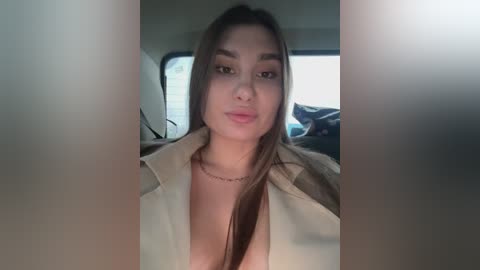 Media: A video of a young woman with long dark hair and light skin, wearing an open beige shirt, revealing cleavage. She is in a car with a blurred background.