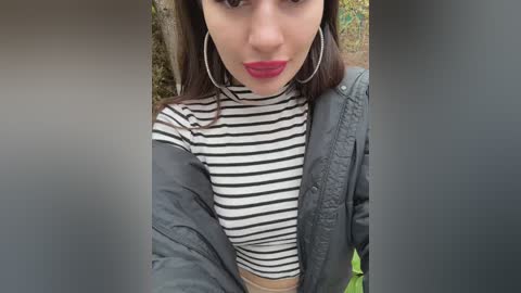 Media: Video of a young woman with fair skin, dark hair, red lipstick, large hoop earrings, and a grey jacket over a black-and-white striped crop top, taken outdoors.