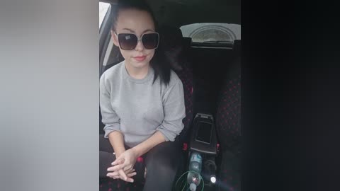 Video of a smiling, fair-skinned woman with long black hair, wearing large sunglasses and a light gray sweater, sitting in a car with patterned seats.