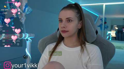 Media: Video of a young woman with fair skin and brown hair in a high ponytail, wearing a white T-shirt, sitting on a gray gaming chair in a dimly lit room.