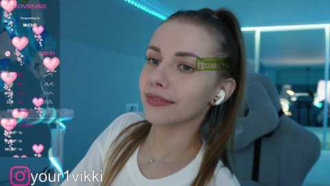 Media: Video of a fair-skinned, young woman with long brown hair in a high ponytail, wearing white headphones and a white top, sitting in a grey gaming chair, in a dimly-lit, modern gaming room.