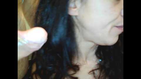 Video of a close-up of a woman with curly black hair, wearing a silver necklace, and a man's hand caressing her shoulder, in a dimly lit room.