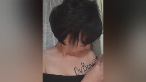 Video of a young woman with short black hair, wearing a black off-shoulder top, holding a tattoo gun, with \"u/Boxxuu\" written on her chest.