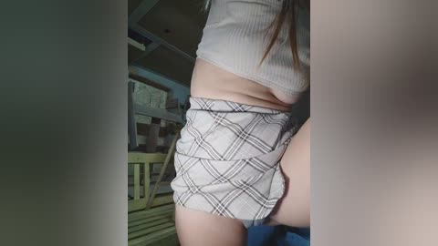 Video of a light-skinned woman wearing a cropped white ribbed crop top and plaid shorts, standing in a dimly lit room with a wooden crib and blue sheets in the background.