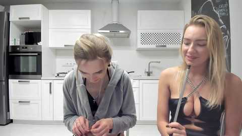 Media: Video of a blonde woman in a revealing black dress with a plunging neckline, applying makeup, and a woman in a grey hoodie adjusting her glasses in a modern, white kitchen.