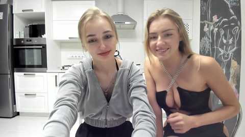 Media: Video of two smiling, blonde, fair-skinned women in a modern kitchen with white cabinets and black appliances. One wears a grey hoodie, the other a black dress with a plunging neckline.