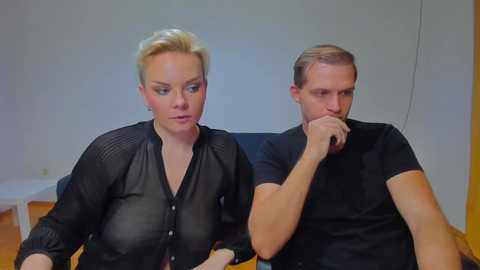 Video of a blonde woman in a black blouse and a man in a black shirt, both seated on a couch with wooden floor, both deep in thought.