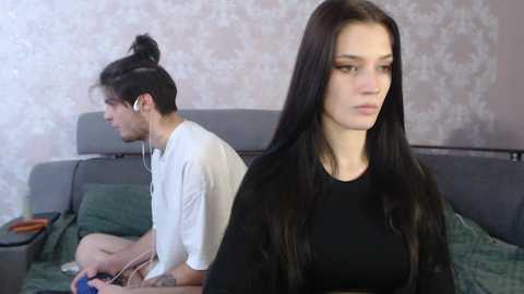 Media: Video of a young woman with long black hair, wearing a black top, sitting on a couch with a patterned wall behind her. A man with a white shirt and a bun hairstyle is listening to music, sitting behind her.