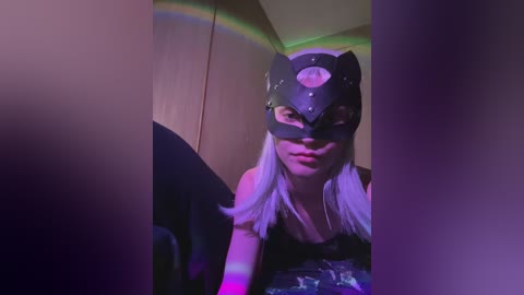 Video of a woman in a black cat mask with a moon design, silver hair, wearing a black top, indoors with purple and green lighting.