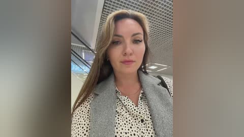 Video of a fair-skinned woman with long, straight blonde hair, wearing a gray blazer over a white blouse with black polka dots. She stands in an indoor setting with a modern ceiling and a staircase in the background.