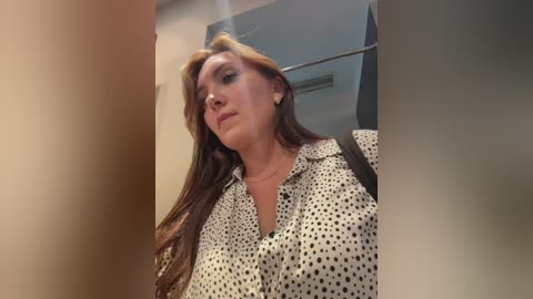 A video of a middle-aged woman with long, wavy brown hair, wearing a polka-dot blouse, standing in a modern, minimalist bathroom with a large mirror reflecting the space.