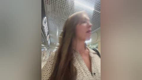A video shows a blonde woman with long, straight hair, wearing a beige, polka-dot sweater, captured in a dimly lit indoor setting with a grid ceiling and blurred signage.