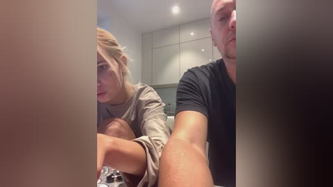 Media: Video of a woman with blonde hair and a man with short hair, both wearing casual clothes, sitting in a modern kitchen with white cabinets and recessed lighting.