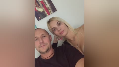 Media: Video of a smiling, light-skinned, blonde woman with medium-length hair and a man with a bald head and beard, wearing black shirts, in a cozy, indoor setting with a colorful, abstract painting on the wall.