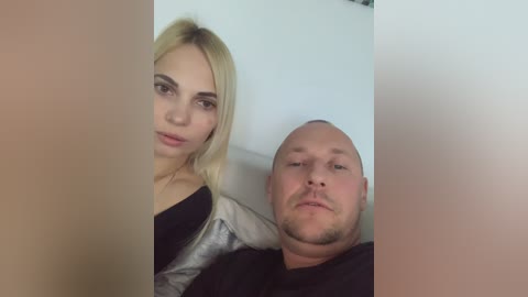 Media: A video of a blonde woman and a bald man, both wearing black shirts, sitting closely together on a couch with a white wall behind them.