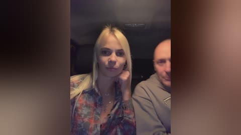 Media: Video of a blonde woman with fair skin, wearing a colorful plaid shirt, leaning on her hand, sitting next to a bald man in a gray hoodie in a dimly lit car.