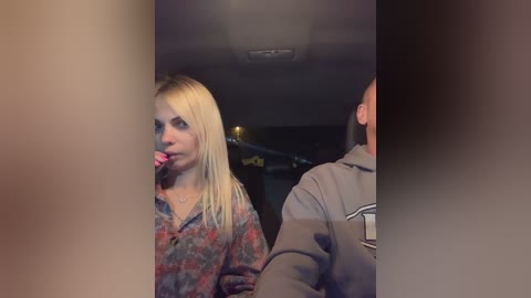 Media: Video of a blonde woman in a colorful patterned shirt, with a lollipop in her mouth, seated in a dimly lit car next to a man in a grey jacket.