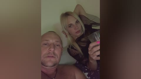 Media: Video of a bald, shirtless, light-skinned man and a blonde, pale-skinned woman with long hair, wearing black lingerie, taking a selfie in a dimly lit room.