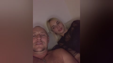 Media: Video of a middle-aged bald man and a young blonde woman in a dimly lit room, both smiling. The man is shirtless with a necklace, and the woman wears a black lace top.