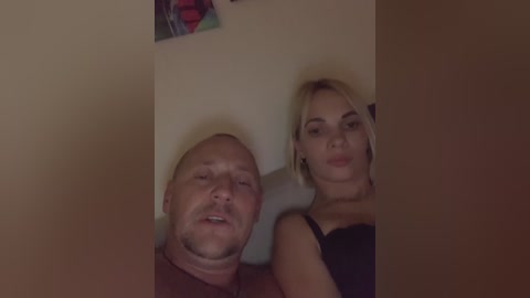 Media: Video of a bald, muscular white man and a blonde woman with a fair complexion and large earrings, both in black tops, leaning against a beige wall.