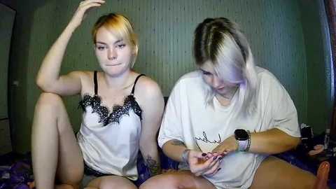 Media: Video of two young women with short hair, one blonde and one dark, wearing casual shirts and sitting on a blue carpet in a dimly-lit room.