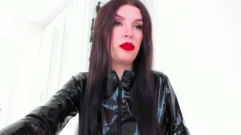 A video of a pale-skinned woman with long, straight dark hair, wearing bold red lipstick and a shiny black leather jacket. The background is a white-walled room with a door.