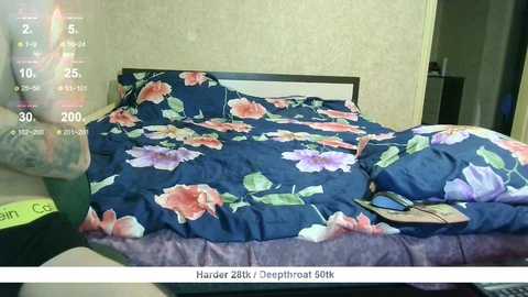 Video of a messy bedroom with a dark blue floral bedspread, scattered clothes, and a partially visible person in the corner. The room has light beige walls and a closed door.