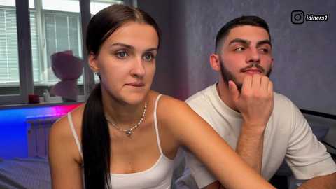 A video of a young, attractive couple with dark hair, seated indoors. The woman, in a white tank top, rests her head on the man's shoulder. Background includes a window, blue and red lights, and a stuffed animal.