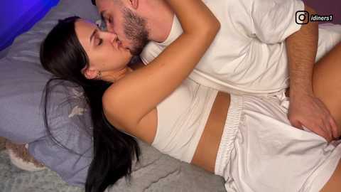 Video of a couple in bed, a brunette woman in a white tank top and shorts, and a bearded man in a white shirt and shorts, kissing passionately.
