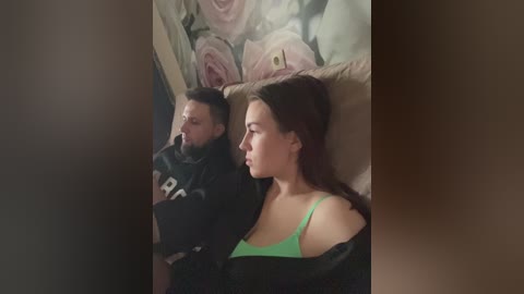 Media: Video of a young couple seated on a couch, man in black hoodie, woman in green bra, both facing right, floral wallpaper in background.