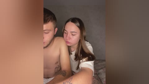A video showing a topless man with tattoos and a woman in a white t-shirt, both with fair skin, cuddling on a bed with a grey blanket.
