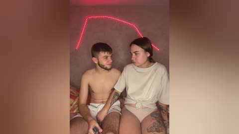 Video of a shirtless young man with a trimmed beard, wearing white shorts, and a tattooed woman in a beige shirt, sitting on a bed with red LED lights on the wall.