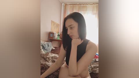 Video of a slender, nude, dark-haired woman with fair skin sitting on a bed in a dimly lit room. The background features a cluttered desk and a window with orange curtains.