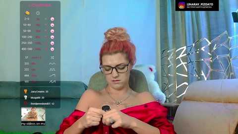 A video of a pale, red-haired woman with glasses, wearing a red satin robe, sitting on a beige couch, fiddling with a black device. Background includes a green couch and a large, white, abstract sculpture.