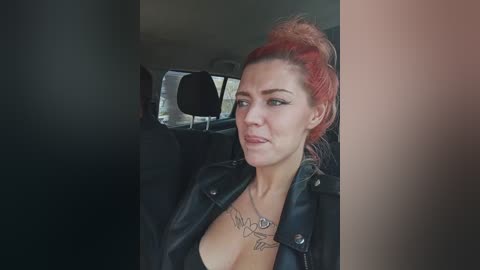 Video of a fair-skinned woman with red hair in a bun, wearing a black leather jacket and revealing a tattoo beneath her cleavage. She's sitting in a car, with a blurry window background.