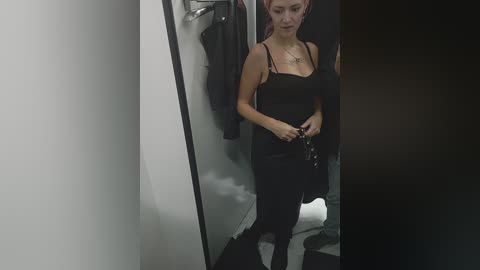 A video of a woman with fair skin and red hair in a black tank top and pants, holding keys, standing in a dimly lit dressing room with a mirror.