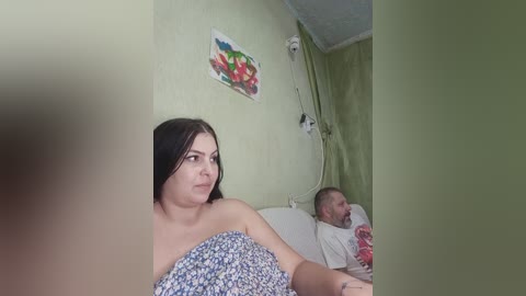 Video of a pale-skinned woman with long black hair, wearing a blue floral dress, sitting next to a middle-aged man with a beard, both in a dimly lit hospital room with green walls and a colorful painting.