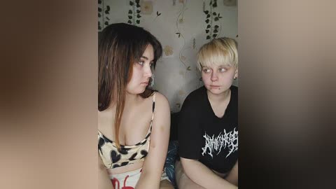 Media: Video of two young women in casual attire; one with long brown hair in a leopard-print top, the other with short blonde hair in a black Slayer t-shirt. Background features a floral-patterned wall and hanging vines.