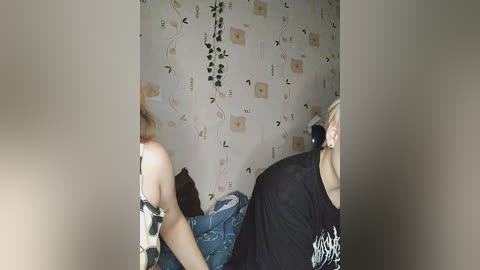 Media: Video of two people in a bedroom with beige walls adorned with floral wallpaper and green ivy vines. One person is in a black t-shirt, and the other in a colorful top.