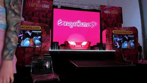 Media: Video of a brightly lit stage with two large, red, brick-patterned screens displaying \"BingaCam\" logos. Two people in colorful attire stand in front of the stage, which has a sleek, black podium.