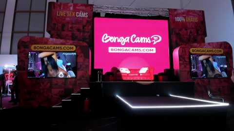 Media: Video of a vibrant, modern stage setup for a live sex cam show featuring a large \"BongaCams\" banner, two monitors displaying performers, and a dimly lit table.