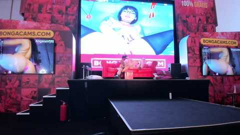 Media: Video of a video game room with a large TV screen showing a woman with blue hair in a video game. Two game consoles with red and black backgrounds flank the screen.