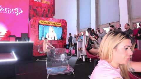 Media: Video of a vibrant red-themed event with a blonde woman in a pink shirt, clear chairs, and a TV screen displaying a woman's face.