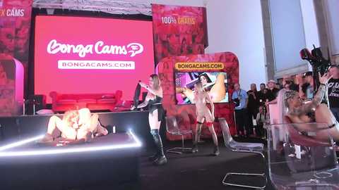 Media: Video of a live performance with two women in transparent plastic suits, one bent over a table, the other on a chair, in a red-lit room with a large screen showing the \"BongaCams\" logo.