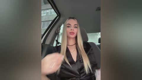 Media: A video of a young woman with long, straight blonde hair, wearing a black satin blouse and choker necklace, seated in a car with a blurry background.