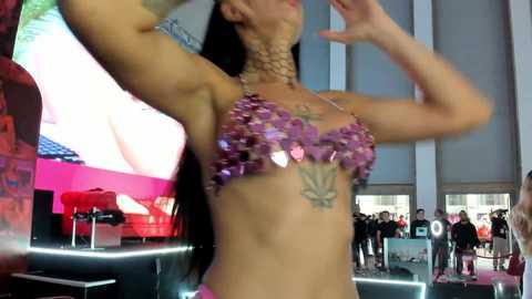 Media: Video of a woman with long black hair wearing a purple sequined bikini top and a beaded choker, posing indoors with a crowd in the background.